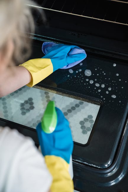 A woman with blonde hair tied back, wearing a white top, yellow and blue cleaning gloves, and a black apron, is engaged in surface cleaning on a white gas stove with four burners. She is using a cloth